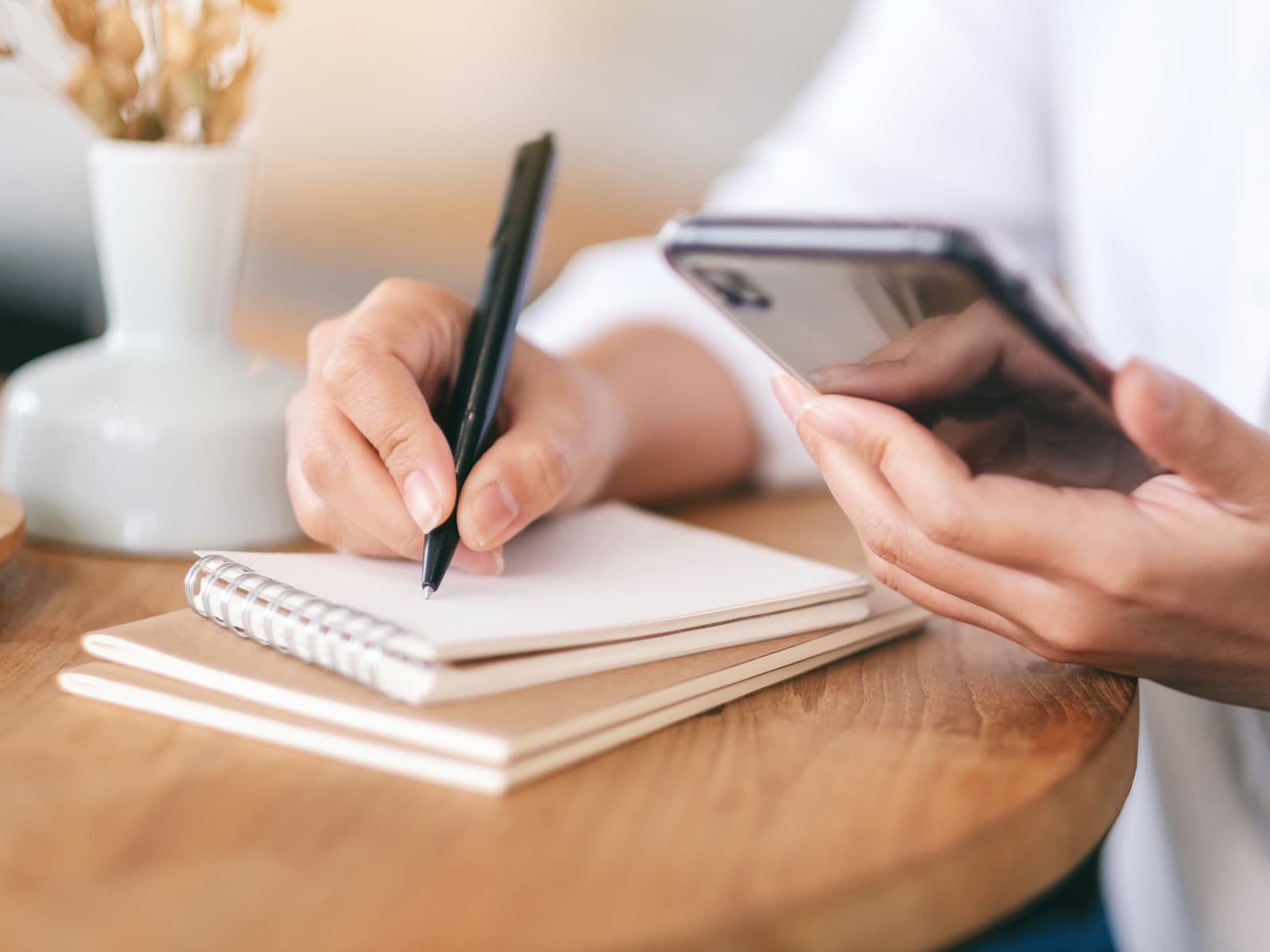 Woman writes in notebook while holding smartphone.