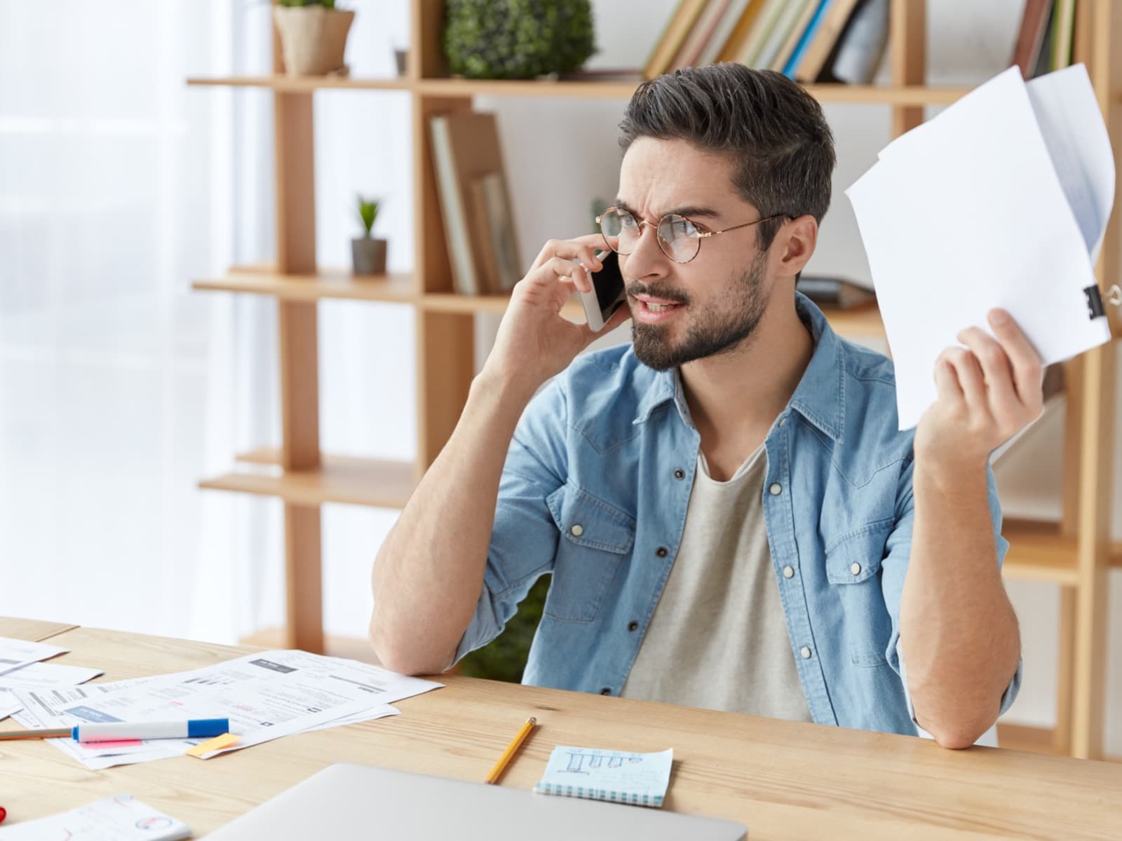 Man on the phone at a desk, holding up a document with a frustrated expression.
